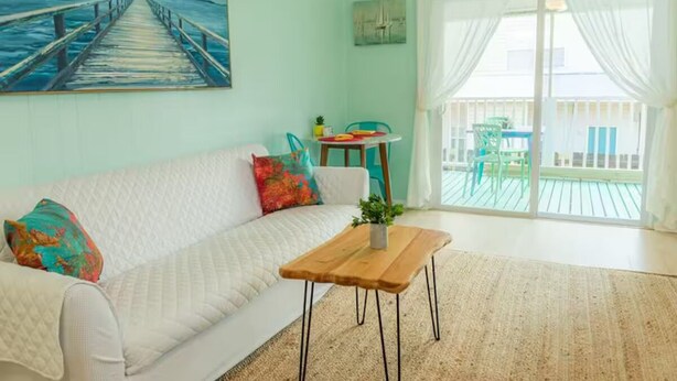 Een woonkamer met een witte bank, kleurrijke kussens, een houten salontafel en een schilderij van een steiger.