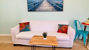 Een woonkamer met een witte bank, kleurrijke kussens, een houten salontafel en een schilderij van een pier boven het water.