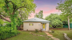 A small, single-story house with a metal roof, surrounded by lush greenery and trees. - Villa 101 · Peaceful River Views · Steps to Water (Glen Rose)