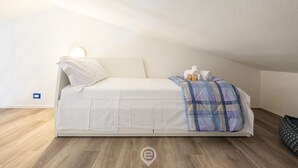 Una cama individual con colchón blanco y manta escocesa, una mesita de noche con lámpara, y una alfombra pequeña sobre suelo de madera en una habitación con paredes blancas y techo inclinado.