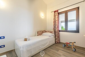 Une petite chambre à coucher, bien rangée, avec un lit simple, une fenêtre avec des rideaux, un plancher en bois et un tricycle pour enfant.
