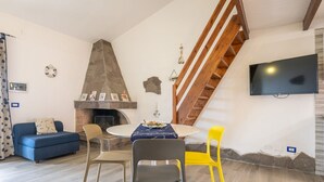 Un salon avec une cheminée en pierre, un canapé bleu, une table à manger avec des chaises, un fauteuil jaune et un téléviseur fixé au mur.