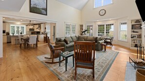 A spacious living area with a high ceiling, wooden flooring, and large windows.