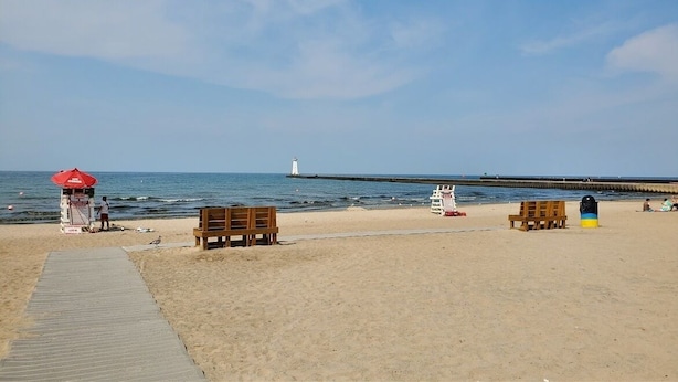 Ein Strand mit einer Holzpromenade, Liegestühlen, einem Rettungsschwimmerstand und einem Leuchtturm in der Ferne.