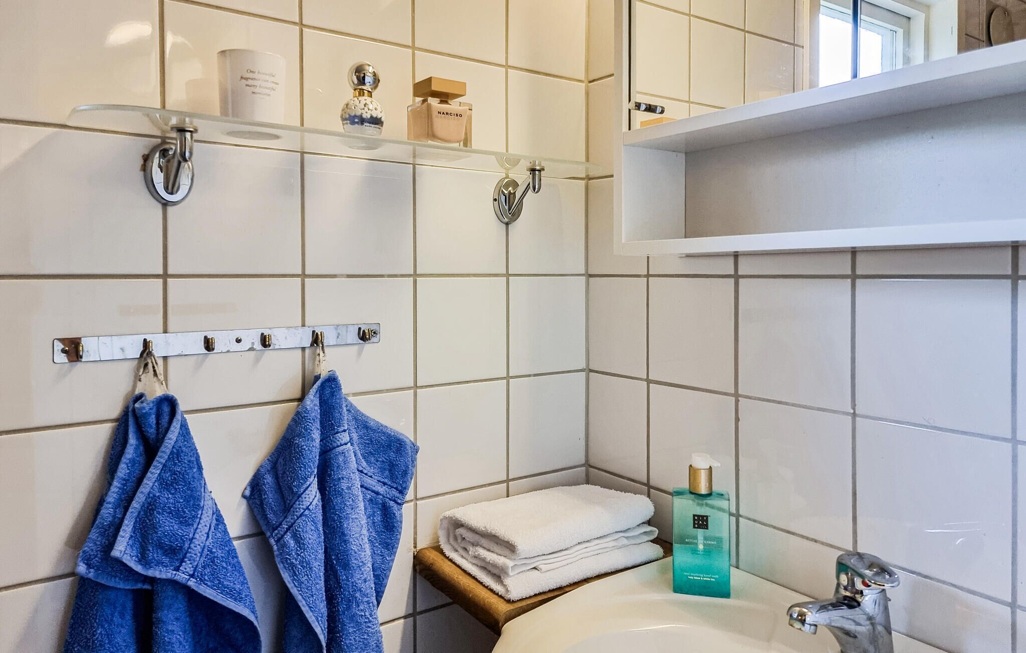 Ein Badezimmer mit weißen Fliesen, einem Waschbecken, einem Spiegel und Regalen mit Pflegeprodukten.