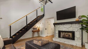 A modern living room with a fireplace, a black leather ottoman, a patterned rug, a staircase with black railings, and a flat-screen TV mounted on the wall. - Cozy Cottage 2 min to WNC AG Center Pet Friendly (Fletcher)