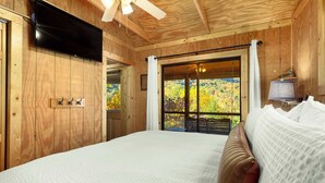 Ein Zimmer mit Holzboden, einem Bett, einem Deckenventilator, einem Fernseher und einem Fenster mit Blick auf Bäume.