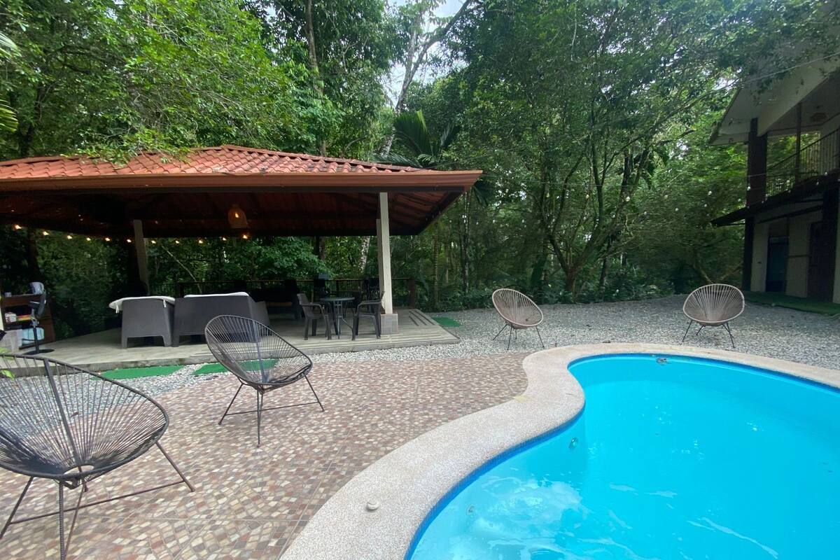A pool area with a tiled deck, lounge chairs, and a covered seating area with tables and chairs.