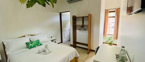 A hotel room with a bed, a desk, a hanging plant, and a window with curtains.