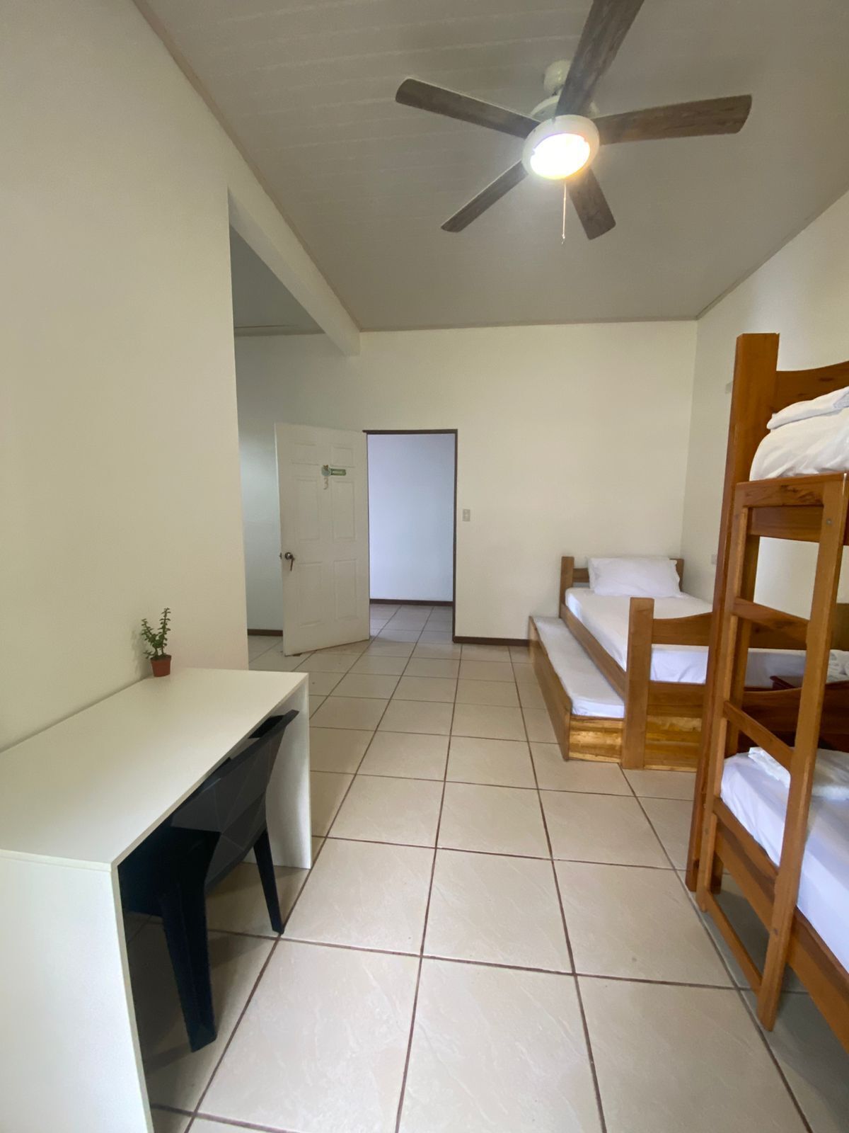 A dormitory room with bunk beds, a desk, a ceiling fan, and a door leading to another room.