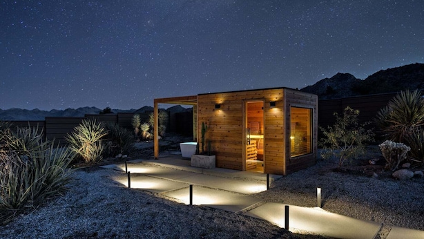A wooden cabin with a patio, surrounded by desert plants, under a starry night sky.