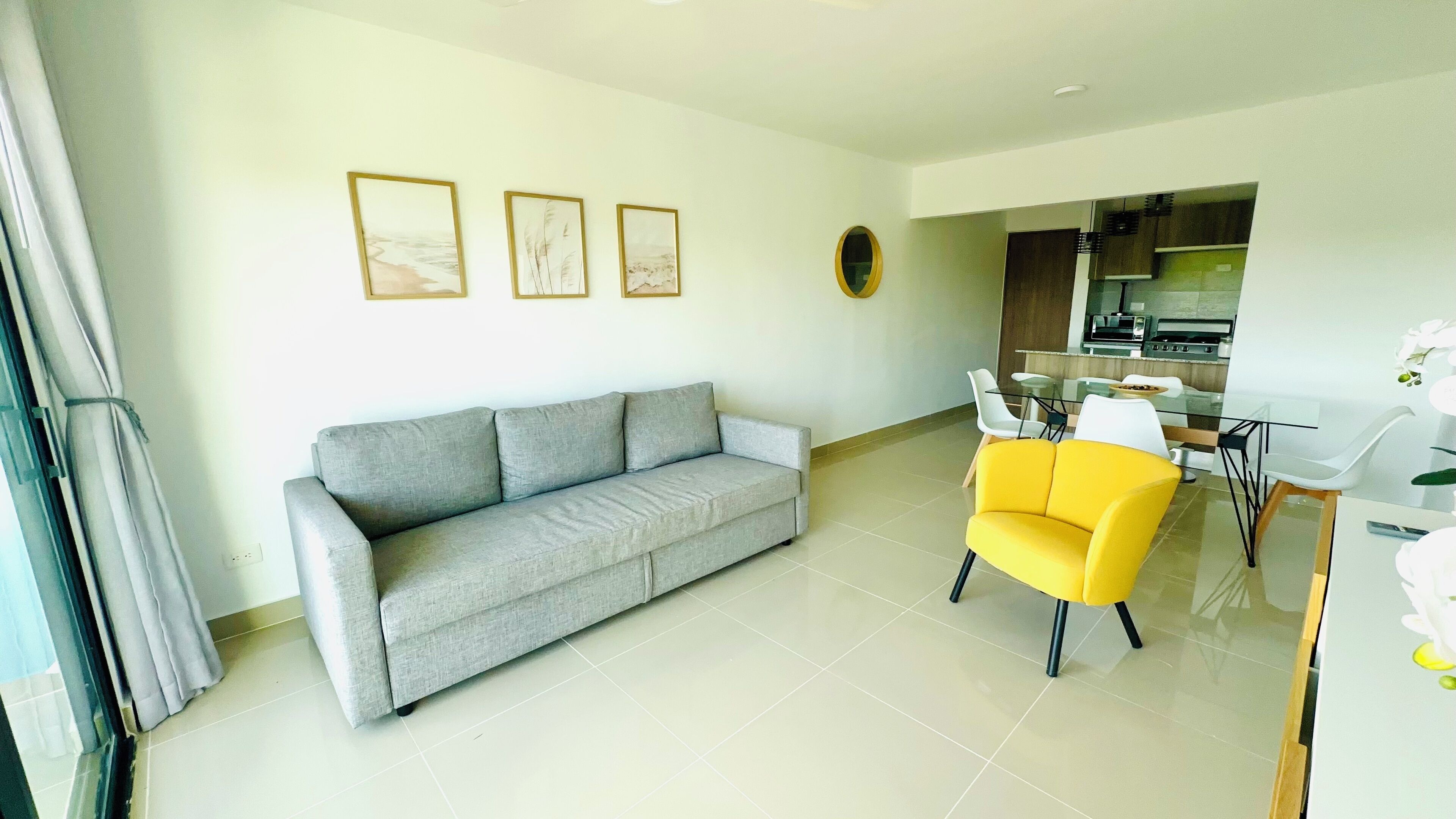 Un salon avec un canapé gris, un fauteuil jaune et un coin repas comprenant une table et des chaises en verre. Trois tableaux encadrés ornent le mur, et on aperçoit une cuisine au fond.
