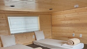 Een kamer met twee bedden, houten wanden, een raam met zonwering en een ventilatie rooster.