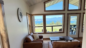 A cozy living room with a brown leather sofa, a wooden coffee table, and a view of a lake and mountains through large windows. - Wild Horse Sunrise at Flathead Lake (Big Arm)