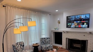 A living room with two floral armchairs, a coffee table, a floor lamp, a fireplace, and a TV mounted on the wall. - Cozy 4-bedroom house in South-Utica! (Utica)