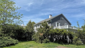 A large, two-story house with a white porch and a grey roof, surrounded by lush greenery.