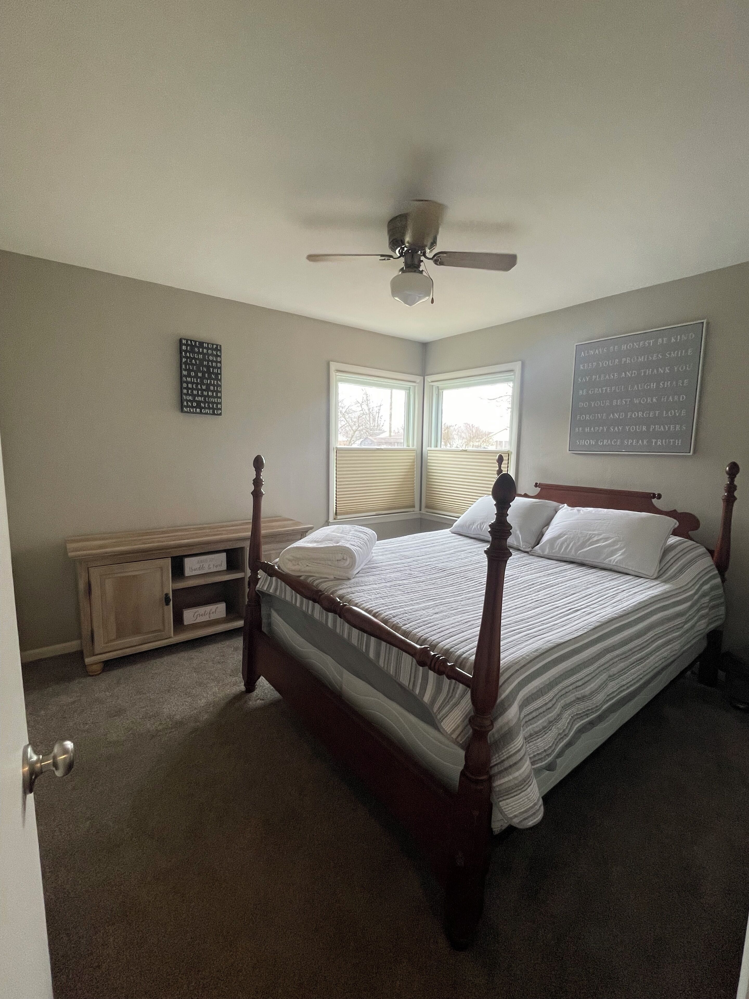 A bedroom with a four-poster bed, a ceiling fan, a window with blinds, and motivational posters on the walls.