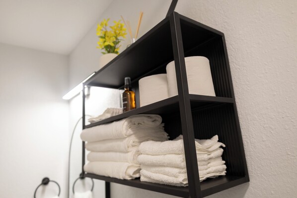 A black shelf with white towels, a bottle, and a roll of toilet paper. - Beautiful, modern furnished 3-bedroom, 2-bathroom cottage near attractions. (Portsmouth)