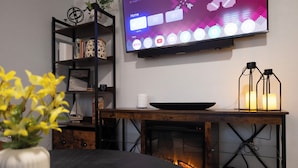 A modern living room with a flat-screen TV mounted on the wall, a wooden coffee table, a bookshelf, and a fireplace. - Beautiful, modern furnished 3-bedroom, 2-bathroom cottage near attractions. (Portsmouth)