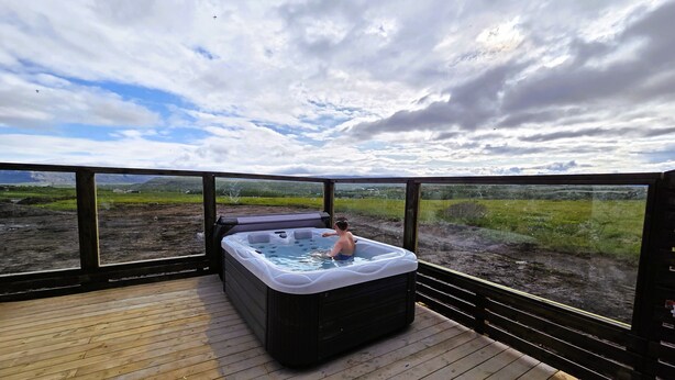 A hot tub on a wooden deck with a view of a grassy landscape and mountains in the distance. - Cosy Northern Ligths Cabin with hot tub and magical views on the Golden Circle (Grímsnes- og Grafningshreppur)