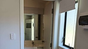 Un pasillo estrecho con una puerta que da a un baño. El baño tiene un lavamanos, un espejo y una ventana con un teléfono.