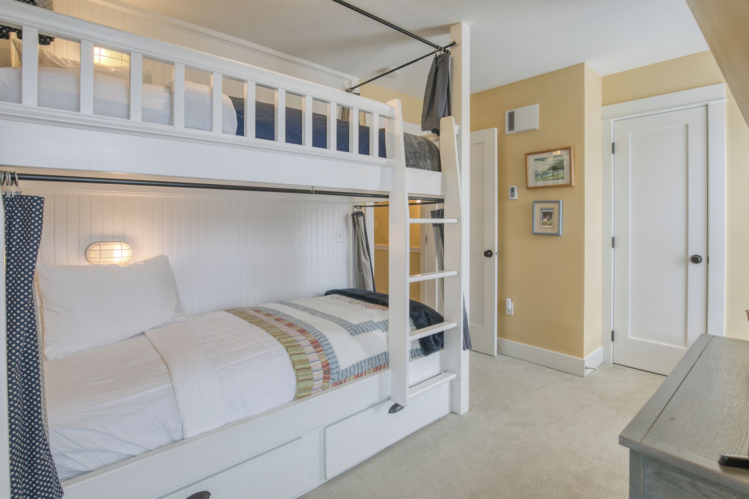 A bunk bed with a ladder, a bedside table, and a door in a room with beige walls and a carpeted floor.