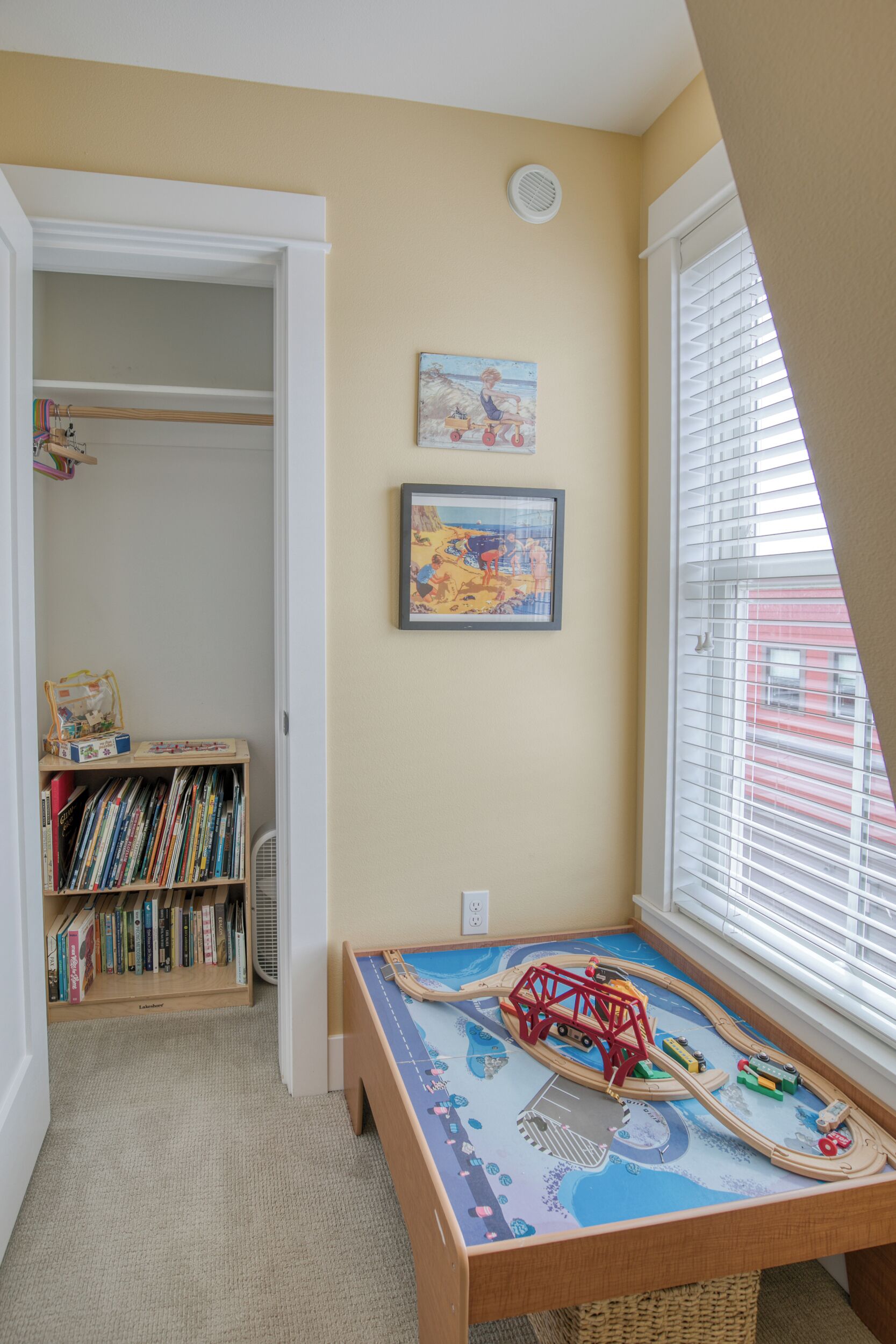 A child's play area with a train set on a table, a bookshelf with books, and two framed pictures on the wall.