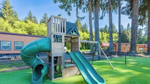 A playground with a slide and climbing structure, surrounded by trees and a building in the background. - Rainforest Retreat Tiny Home: Fishing Cabin (Forks)