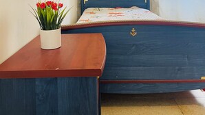 A bedroom with a bed, a wooden nightstand, a potted plant, and a window with a sheer curtain.