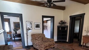 Amplia sala de estar con pisos de madera, un sofá grande con estampado floral, un otoman tapizado y un ventilador de techo. Hay un banco de madera y una mesita con un jarrón de flores.