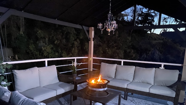 A covered outdoor seating area with a fire feature, white cushioned sofas, and a wicker chair.