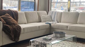 A living room with a beige sectional sofa, a glass coffee table with magazines, and a view of boats through large windows.