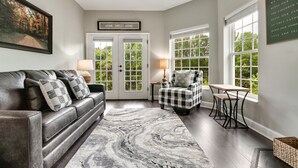 A living room with a dark leather sofa, a checkered armchair, a marble-patterned rug, and a view of a forest through large windows.