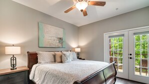 A bedroom with a bed, nightstands, a ceiling fan, a painting, and a door leading to an outdoor area.