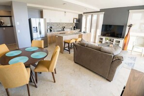 A modern kitchen with a central island, a dining area with a round table and chairs, a living room with a sofa and a TV, and a large window providing natural light.