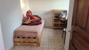 A small, narrow room with a bed, a wooden shelf, and a patterned floor.