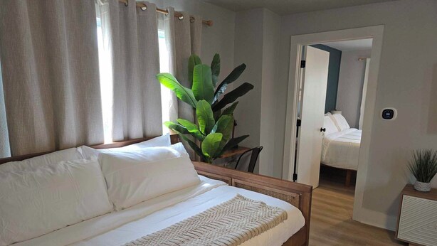 Un dormitorio con cama de madera, ropa de cama blanca, una planta verde grande y vistas a otra habitación.