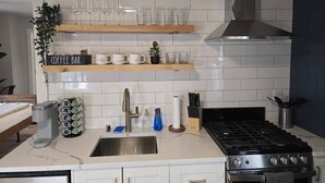 Una cocina con gabinetes blancos, un lavamanos de acero inoxidable, una cafetera, una estufa y estantes con cristalería y un letrero que dice 'Bar de café'.