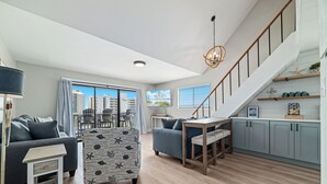 A modern living room with a grey sofa, patterned armchair, and a dining area with a table and chairs. A staircase with a wooden railing leads to a kitchen area with light blue cabinets and a small island. - Skip the packed high-rises!•
Steps to Beach • Family Ready • 
Gulf View 2BR-2BA (Orange Beach)