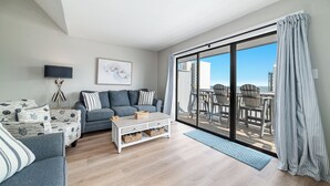 A modern living room with a grey sofa, a white coffee table, and a large sliding glass door leading to an outdoor deck with chairs. - Skip the packed high-rises!•
Steps to Beach • Family Ready • 
Gulf View 2BR-2BA (Orange Beach)