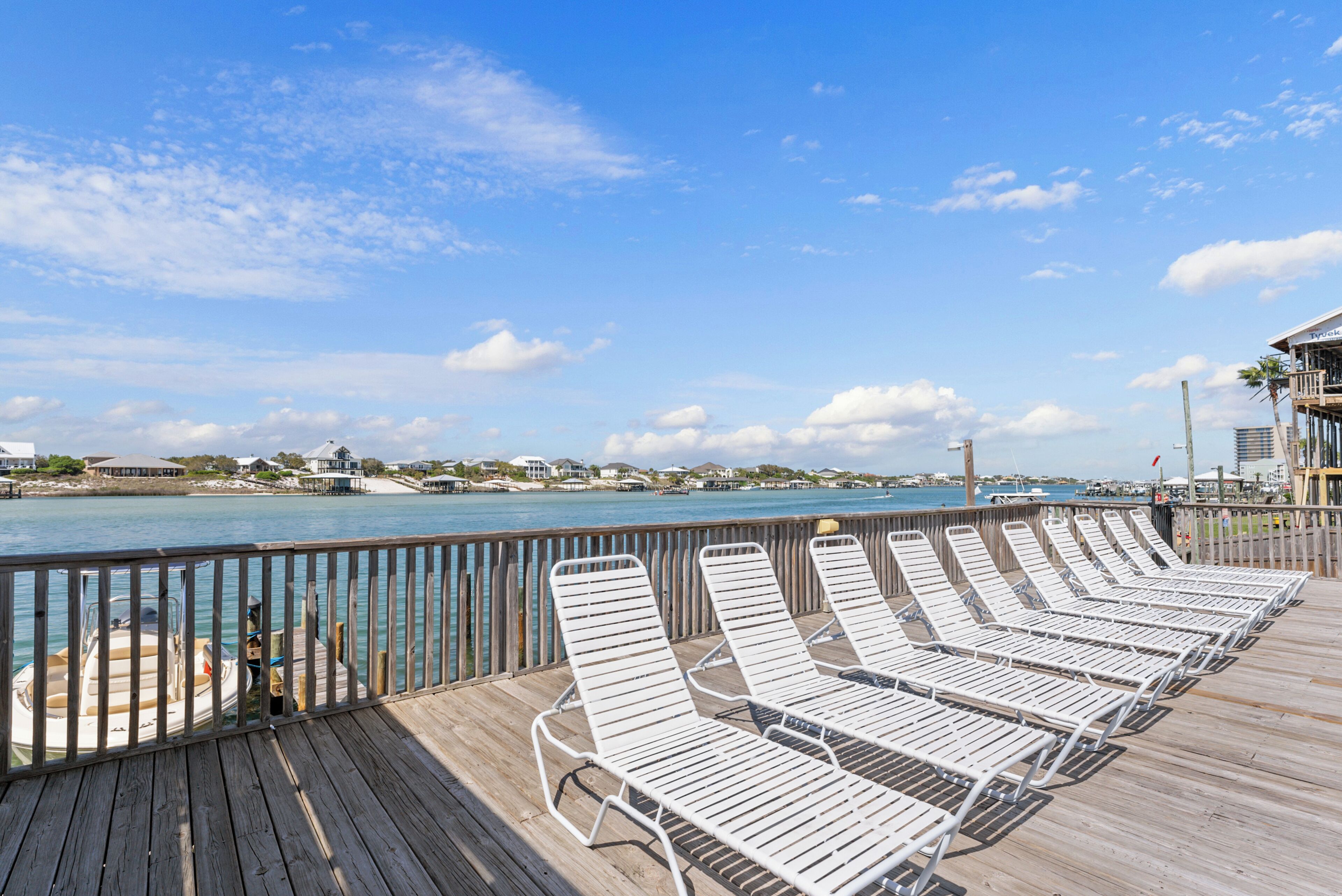 A wooden deck with white metal sun loungers facing a waterfront.