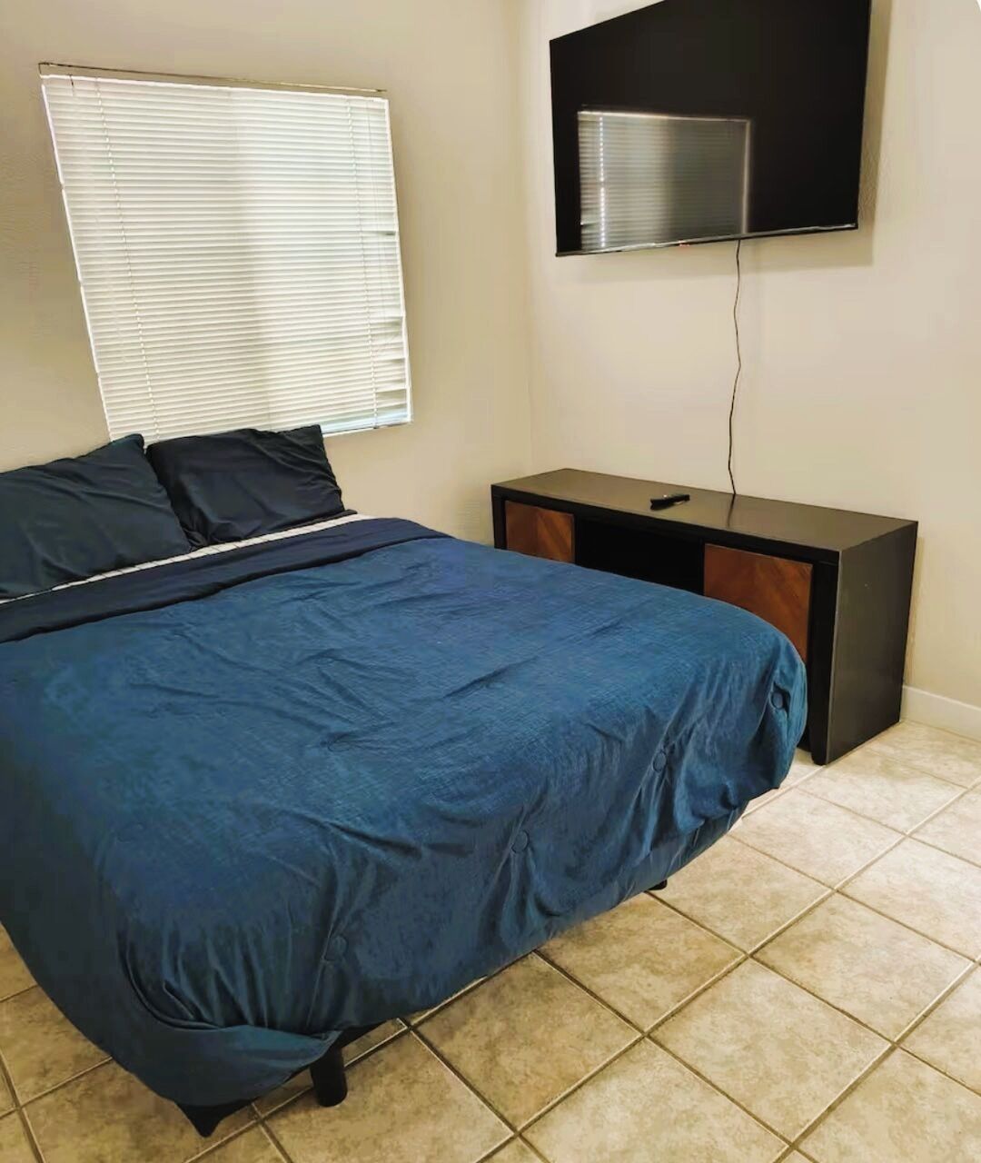A bedroom with a bed, a nightstand, a TV mounted on the wall, and a window with blinds.