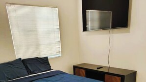 A bedroom with a bed, a nightstand, a TV mounted on the wall, and a window with blinds. - Style & Convenience Near PHX Ask for LongTerm Rate (Tempe)