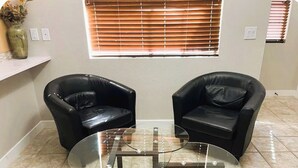 A room with two black leather chairs, a glass coffee table, a vase with dried flowers, and a window with blinds. - Style & Convenience Near PHX Ask for LongTerm Rate (Tempe)