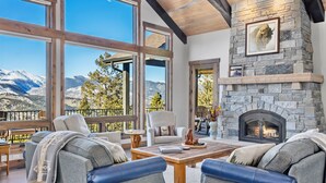 Un salon chaleureux avec une cheminée en pierre, de grandes fenêtres offrant une vue sur les montagnes et des sièges confortables.