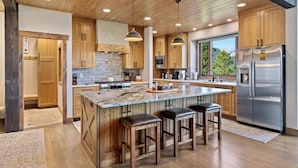 Une cuisine moderne avec des meubles en bois, un grand îlot central doté de tabourets, des appareils électroménagers en acier inoxydable et une vue sur la verdure environnante.