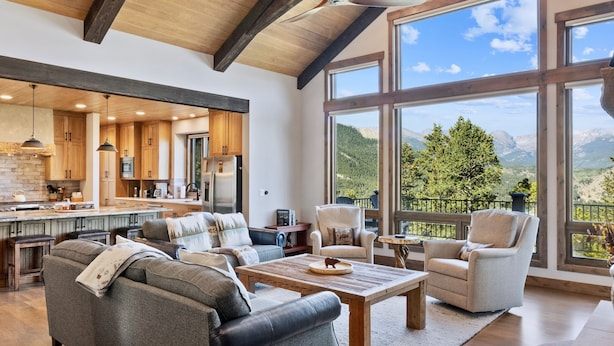Un vaste espace de vie doté d’un plafond haut, de poutres en bois, d’une grande fenêtre offrant une vue sur les montagnes et d’une cuisine moderne en arrière-plan.