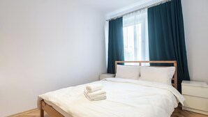 A neatly made bed with white linens and a rolled towel, a wooden headboard, and a nightstand with a white lamp.