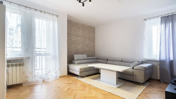 A modern living room with a herringbone wooden floor, a grey sectional sofa, a white coffee table, and a brick accent wall.