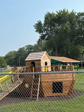 A wooden playhouse with a ramp and a slide, enclosed by a chain-link fence.